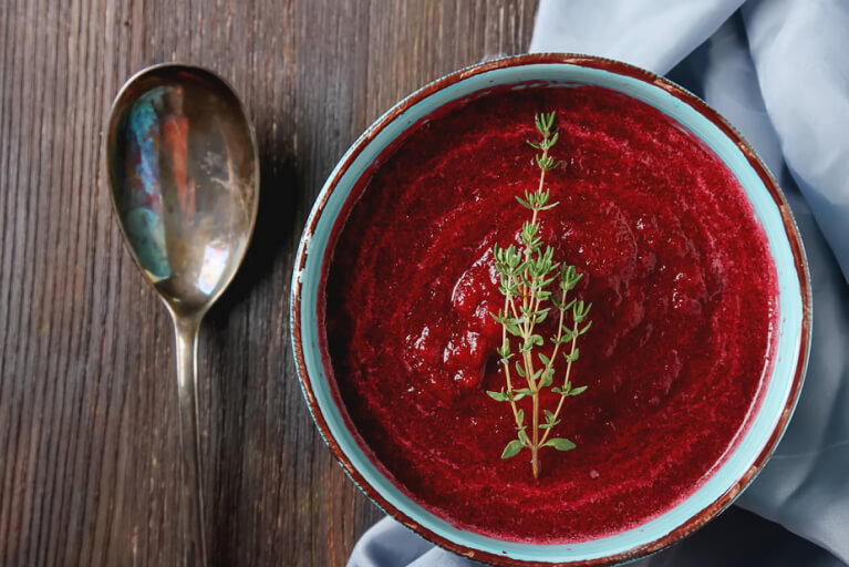 Vue supérieure. Crème de betteraves dans un bol bleu déposé sur un tissu bleu clair, avec une cuillère à soupe placée à gauche.