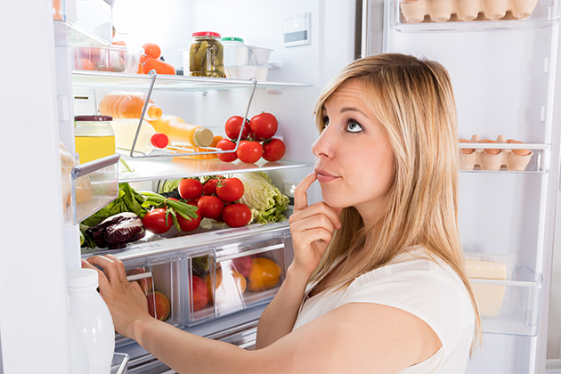 Une femme regarde dans son réfrigérateur, le regard perplexe