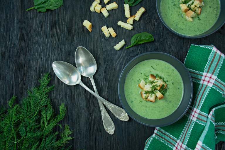 Deux cuillères et deux bols à soupe foncés avec un potage vert de poireaux avec aneth et croûtons en décoration sur le dessus.