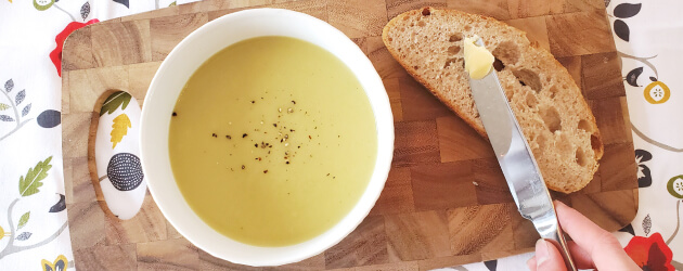 Vus de haut, un bol de soupe avec potage crémeux, tranche de pain et couteau à beurre déposés sur une planche de bois.