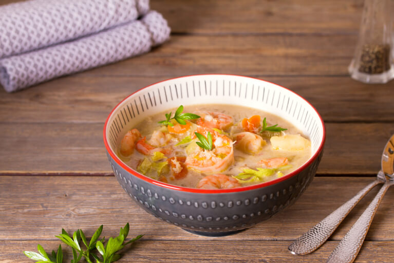 Des cuillères à soupe et un bol avec un bouillon clair, poireaux et crevettes sur une table en bois.