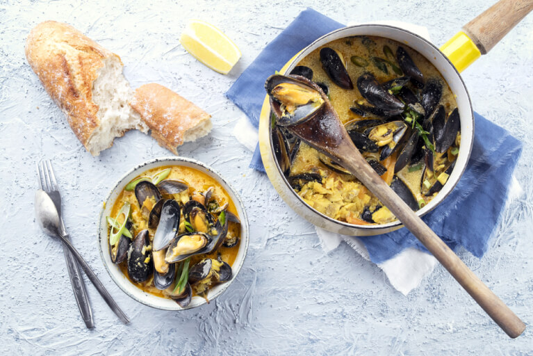 Un bol et un chaudron jaune remplis d'un velouté aux moules et poireaux, avec une cuillère de service en bois.